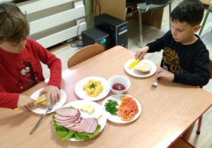 Chłopcy smarują masłem bułeczki. Na stoliku stoją talerze z wędliną, warzywami, jajecznicą, salaterka z dżemem
